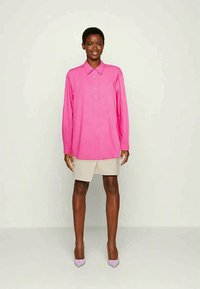 Mulher vestindo uma camisa de botão de manga longa rosa brilhante, calções bege até ao joelho e saltos baixos roxos claros, de pé contra um fundo plain.