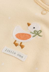 Kremna tkanina z vezeno belo in oranžno raco, ki drži zeleno pentljo, z majhno etiketo z napisom "little one" pod raco.