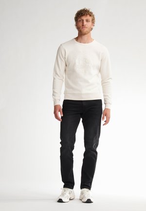 Mann mit lockigem Haar, der ein weißes langärmliges Hemd, schwarze Jeans und weiße Turnschuhe trägt und vor einem einfarbigen weißen Hintergrund steht.