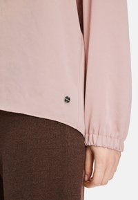 Rosa Bluse aus leichtem Stoff mit gerafften Ärmeln, mit einem silbernen Logo-Detail am Saum. Braune strukturierte Hose teilweise sichtbar.