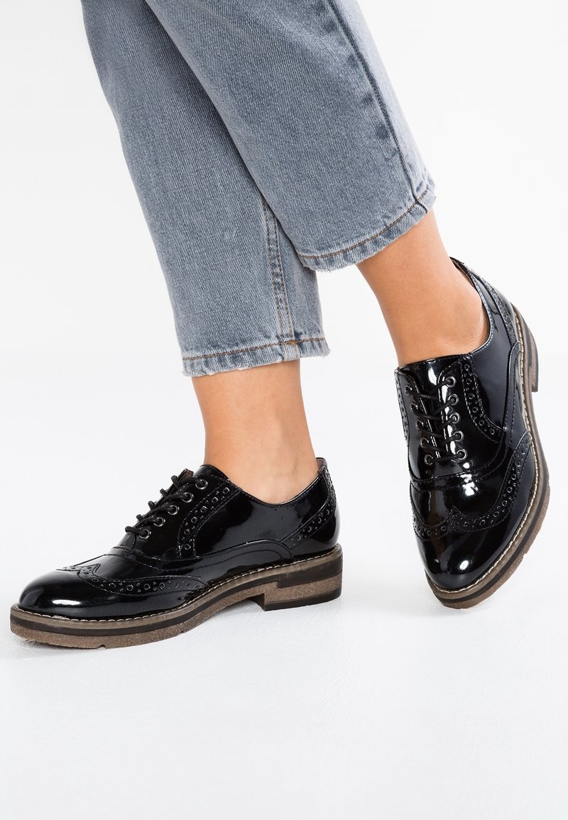 Personne portant un jean court bleu clair et des chaussures brogues en cuir noir brillant avec des semelles épaisses marron.