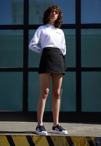 Chemise blanche à manches longues avec des accents imprimés, jupe portefeuille noire et baskets noires avec des rayures blanches, se tenant devant un arrière-plan en verre.