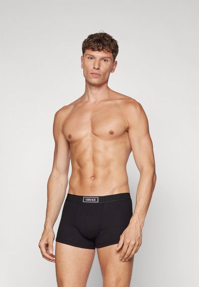 Boxer-briefs noirs en tissu extensible, dotés d'une ceinture avec un logo "VERSACE" subtil. Le modèle pose avec une expression neutre.
