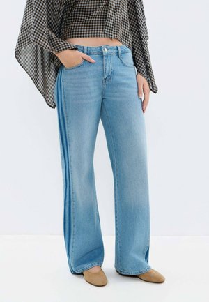 Personne portant un jean large bleu clair avec des bandes latérales foncées, un haut à manches longues marron à carreaux, et des chaussures beiges à bouts pointus, debout avec une main dans la poche.