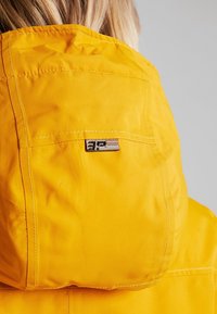 Veste imperméable jaune avec une finition texturée, dotée d'une capuche et d'une petite étiquette de marque au dos. Les coutures surpiquées sont visibles.