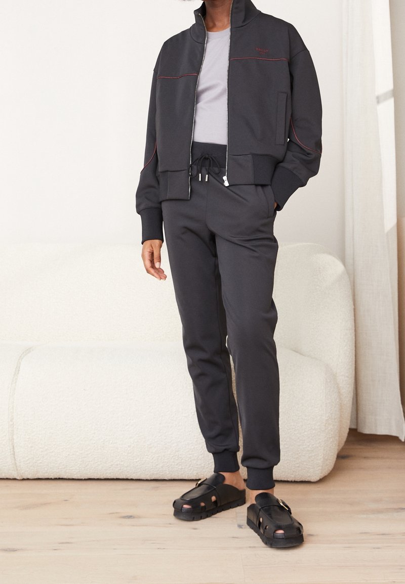 Giacca nera con zip e cuciture rosse, abbinata a pantaloni joggers coordinati e scarpe slip-on nere. Tessuto testurizzato e design aderente.