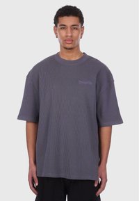 T-shirt gris surdimensionné avec des manches courtes, texture côtelée, et un petit logo brodé sur la poitrine. Fabriqué à partir d'un tissu léger.