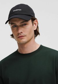 Fekete baseball sapka "lululemon" logóval, sötétzöld pulóverrel párosítva, amelyen kerek nyak és bordás mandzsetta található.