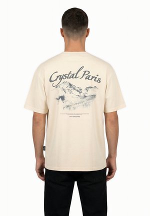 Uomo di spalle che indossa una maglietta beige con la scritta "Crystal Paris" e un'illustrazione di montagna sul retro, abbinata a pantaloni neri.