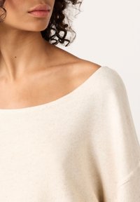 Šviesiai smėlio spalvos megztinis iš mezgimo siūlų, su plačiu valtiniu iškirpimu, tekstūruota medžiaga ir nuleistais pečiais. Medžiaga atrodo minkšta, su subtiliu taškuotu raštu.