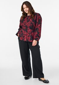 Kvindelig med langt, bølget hår iført en rød og sort blomsterbluse, sorte brede bukser og sorte flade sko med nitter på en hvid baggrund.