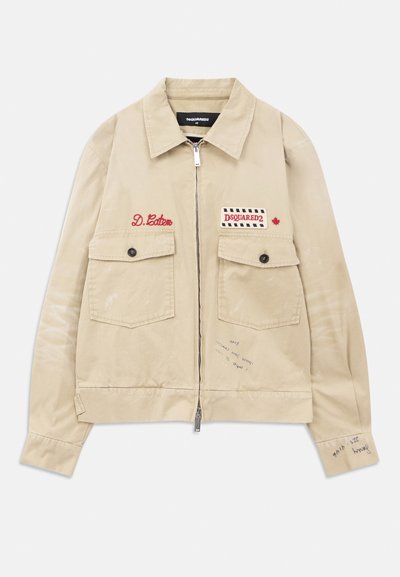 Chaqueta beige con cremallera y dos bolsillos frontales abotonados, parche rojo bordado "D. Caten," "DSQUARED2" y detalles de texto manuscrito en la parte delantera y en la manga.