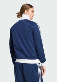 Tamsiai mėlynas sportinis džemperis su aukštu apkakle, baltais akcentais ir trimis baltomis juostelėmis ant rankovių. Raštuotas audinys, prigludęs sijonas.