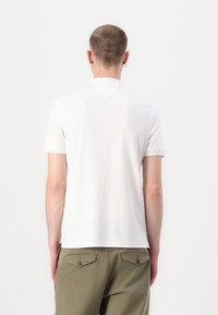 Korte mouwen witte polo met een zachte textuur, geribbelde kraag en een klein driehoekig detail op de rug, gecombineerd met groene broek.