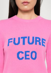 Frau mit einem leuchtend pinken Pullover, auf dem in blauer Schrift "FUTURE CEO" auf der Brust zentriert ist, vor einem schlichten Hintergrund.