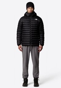 Svart boblejakke med glidelås foran og hette, med horisontal quilting. Brukt med grå joggebukser og svarte tursko.