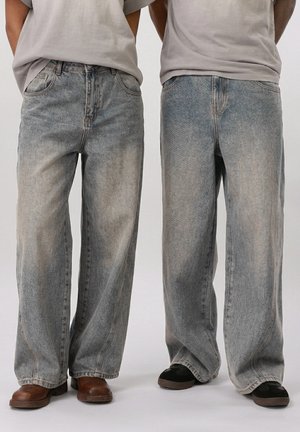To personer iført lyse, løstsiddende jeans med falmet detaljering, kombineret med afslappede brune og sorte sko, stående side om side.