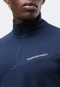 Κοντινό πλάνο ατόμου που φοράει μπλούζα navy blue με φερμουάρ ως το στήθος και λογότυπο "Peak Performance" στο στήθος.