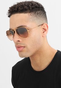 Ochelari de soare tip aviator cu lentile maro închis, ramă din metal aurit și detalii fine pe brațe, purtați cu un tricou negru cu guler rotund.