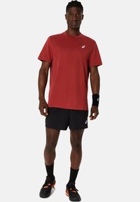 Červené sportovní tričko s krátkým rukávem a logem Asics, spárované s černými šortkami a černými ponožkami do kotníků. Černé a oranžové sportovní boty.