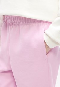 Κοντινό ενός χεριού που βρίσκεται στην τσέπη ροζ υφασμάτινου παντελονιού με ελαστική μέση, ενώ φοράει λευκό μακρυμάνικο τοπάκι.