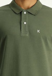 Oliwkowa koszulka polo wykonana z bawełny, posiadająca zapięcie na trzy guziki oraz małe haftowane logo po lewej stronie. Lekki i strukturalny materiał.