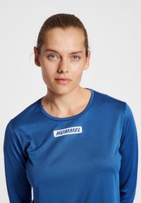 Blå atletisk tröja med lång ärm, rund halsringning och en vit logotyp på bröstet. Tyget ser slätt ut med en lätt glans.