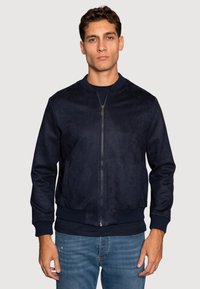 Chaqueta bomber de la marina hecha de suave ante, con cierre de cremallera en el frente, puños y dobladillo acanalados, y dos bolsillos laterales. Llevada sobre una camiseta oscura.