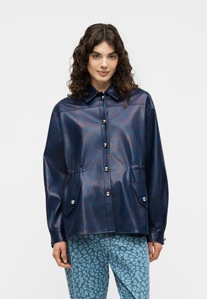 Frau mit lockigem Haar, die eine glänzende marineblaue Jacke mit Knöpfen und Klapptaschen sowie blaue gemusterte Hose trägt, steht vor einem schlichten Hintergrund.