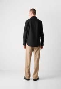 Black long-sleeve shirt with a button-up collar, paired with beige pants, visible seams, and rubber-soled shoes from a rear view.