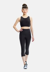 Woman posing in black sleeveless crop top, high-waisted capri leggings, and black-and-white sneakers against a white background.