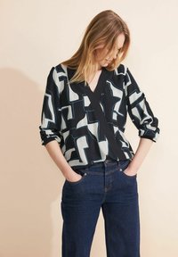 Donna che indossa una blusa a motivi geometrici bianco e nero con le maniche arrotolate e jeans blu scuro, mani nelle tasche, che guarda in basso.
