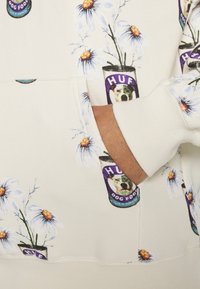 Felpa HUF color crema con stampe di fiori e lattine di cibo per cani. Presenta una tasca frontale e polsini a costine. Tessuto morbido. Design casual.