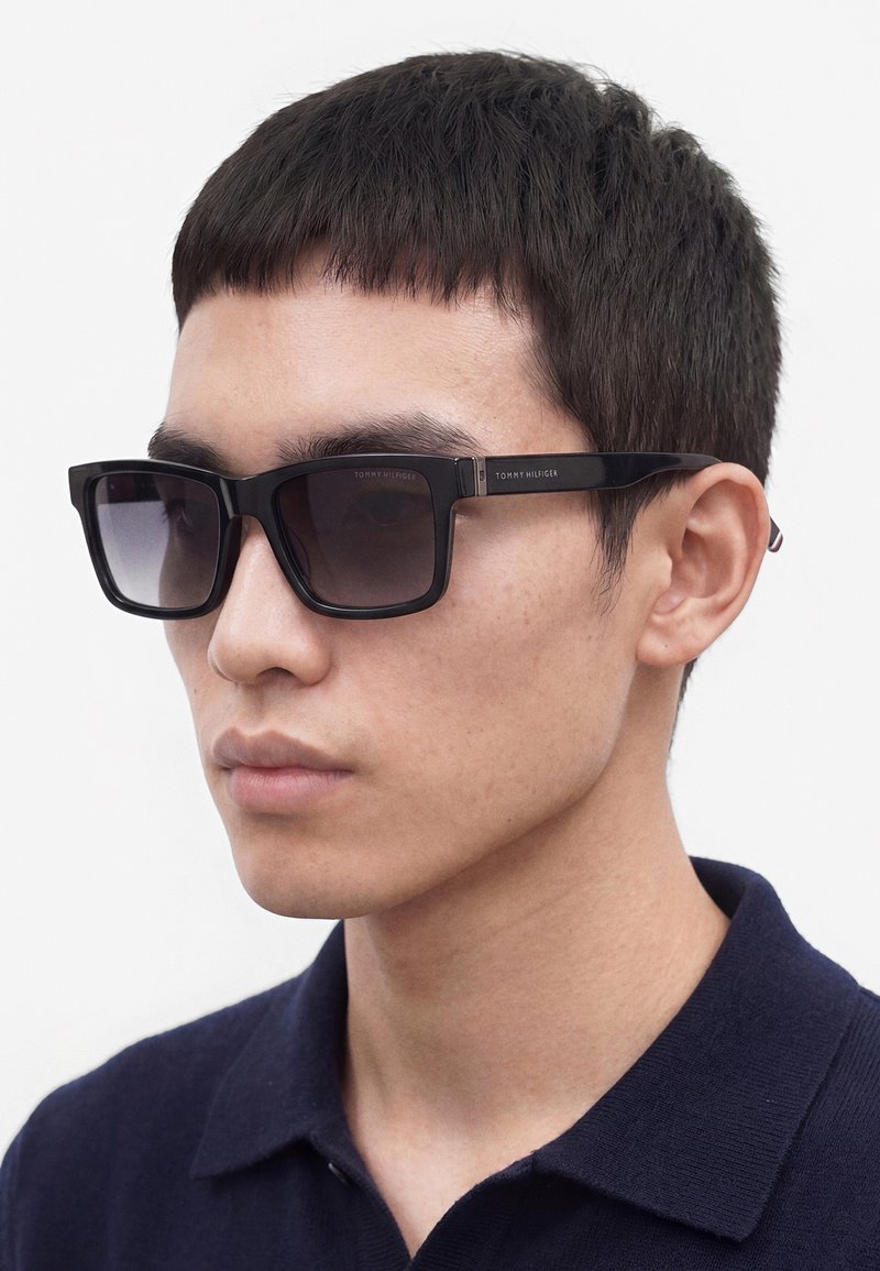 Jeune homme portant des lunettes de soleil noires carrées avec des verres foncés et une chemise bleu marine à col, sur un fond uni.