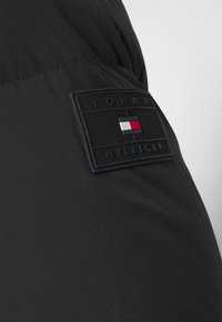 Czarny materiał o gładkiej teksturze z wypukłą prostokątną naszywką. Naszywka prezentuje napis "TOMMY HILFIGER" oraz kolorowe logo flagi.