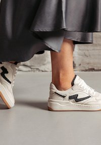 White sneakers with black accents, featuring a textured design, lace-up front, and a gum sole. Part of a person’s foot is visible, wearing a dark skirt.