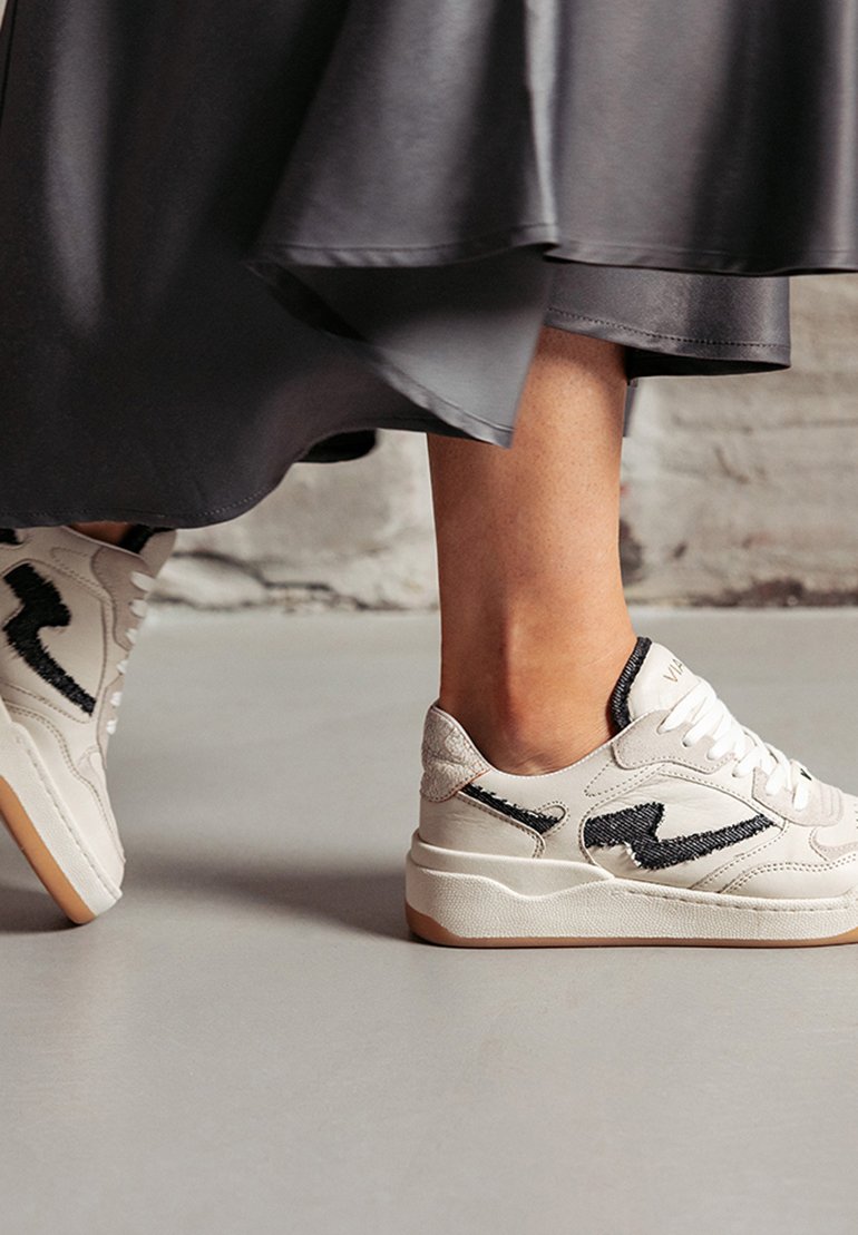 White sneakers with black accents, featuring a textured design, lace-up front, and a gum sole. Part of a person’s foot is visible, wearing a dark skirt.