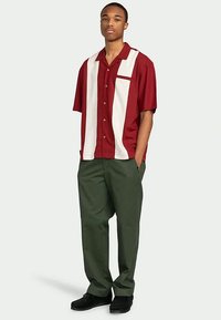 Junger Mann steht in einem kurzärmeligen bordeauxroten und weißen Hemd mit grünen Hosen und schwarzen Schuhen.