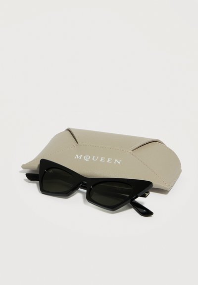 Μαύρα γυαλιά ηλίου με σχήμα γάτας που ξεκουράζονται πάνω σε μπεζ δερμάτινη θήκη με την επιγραφή "McQueen" σε λευκά γράμματα.