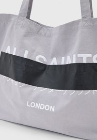 Borsa tote in tela grigia con grande testo bianco "ALL SAINTS" e una striscia orizzontale nera. Caratterizzata da un tessuto resistente e manici lunghi per un facile trasporto.