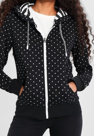 Kvinde iført sort lynlåshættejakke med små hvide prikker, sort-hvidt stribet hættelining, hænder i forlommer.