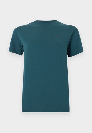 Kortermet t-skjorte i teal med rund hals og liten rød brodert logo på venstre bryst mot en ensfarget bakgrunn.