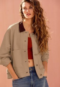 Veste matelassée beige avec un col foncé, grandes poches avant et fermeture à bouton, associée à un crop top rouille et un jean en denim.