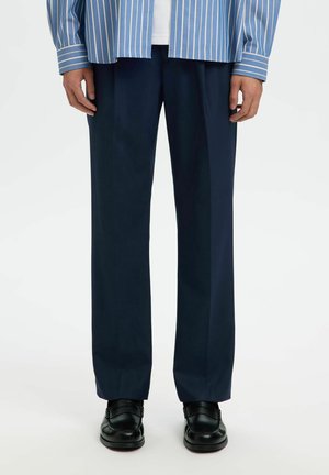 Uomo che indossa pantaloni blu navy a gamba dritta, scarpe di pelle nere, camicia bianca e camicia a righe blu a maniche lunghe, in piedi su sfondo bianco.