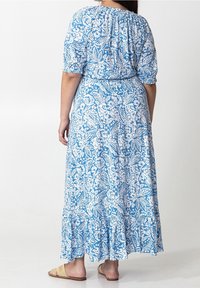 Kvinna som bär en maxi-klänning med blå och vita blommönster, puffärmar och handvävda platta sandaler, stående mot en enkel vit bakgrund.