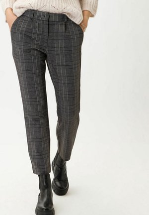 Persoon draagt grijze geruite broek, zwarte leren enkellaarsjes en een crèmekleurige gebreide trui met handen in de zakken.
