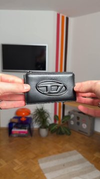 Juoda odinė piniginė, ant kurios yra iškilus ovalo formos logotipas, stačiakampio formos, su užtrauktuku ir lygiu paviršiumi. Rodoma rankomis prieš foną.