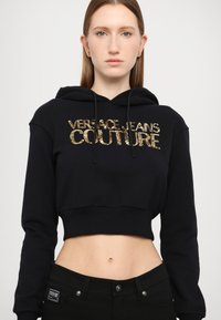 Zwart cropped hoodie met gouden pailletten 'VERSACE JEANS COUTURE' lettering. Voorzien van een trekkoord kap en geribde zoom. Gecombineerd met zwarte jeans.