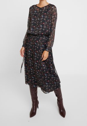 Femme portant une robe midi noire transparente à imprimé floral multicolore et manches longues, associée à des bottes hautes pointues marron foncé.