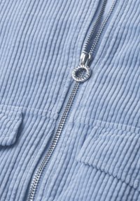 Close-up of silver zipper on light blue ribbed fabric with two rectangular pockets on either side of the zipper.